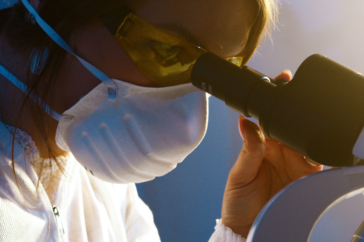 A masked scientist in a white lab coat looking into a microscope 
